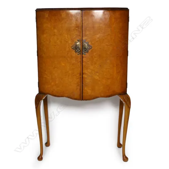 A c.1950 quality figured pale walnut drinks cabinet with the two bow front doors enclosing a mirrored back and base and with two glass shelves. Raised on cabriole legs with acanthus carved knees. 860 x 490 x 1480mm. Provenance: From the collection of Gerry and Valerie Bermel (of Travis Antiques, Wellington 1950's to 1986).
