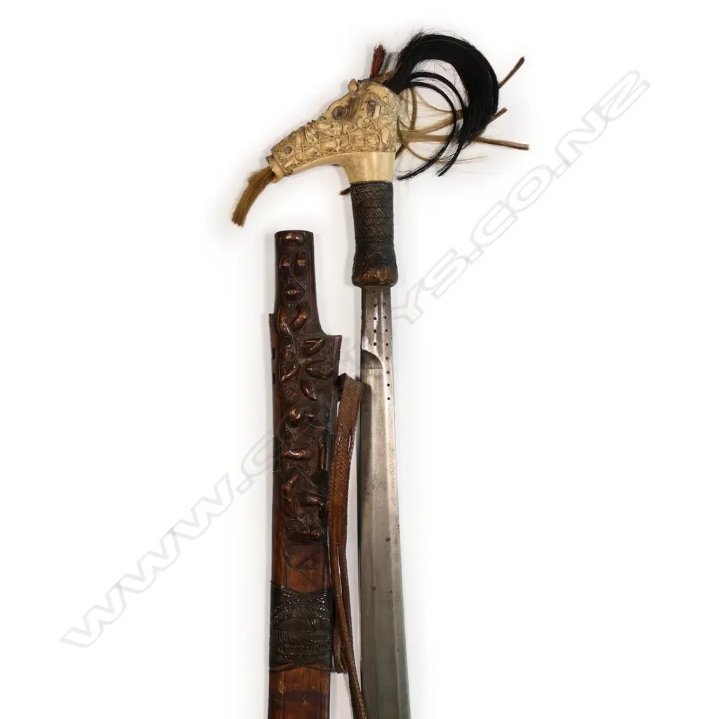 A 19thC Borneo Dayak head-hunter’s sword (Mandau) - Kalimantan region. With plaited fibre lashing to the figure carved wooden scabbard, the carved bone hilt with grotesque animals and scrolling forms and hair tufts highlights. The shaped blade narrowing to the handle. L.700mm. Wooden scabbard split to blade side. Image 1++
