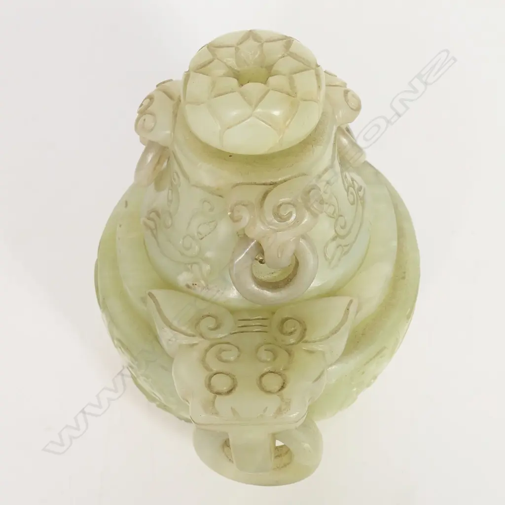 An old Chinese green jade lidded censor - the lid with pronounced flower head finial and three captured ring handles. carved with dragon masks, the body with further conforming masks to the large flat top captured ring handles. The body with opposing stylised dragons and paw feet. L.160mm. Raised on wood stand. H.195mm. Image 1++