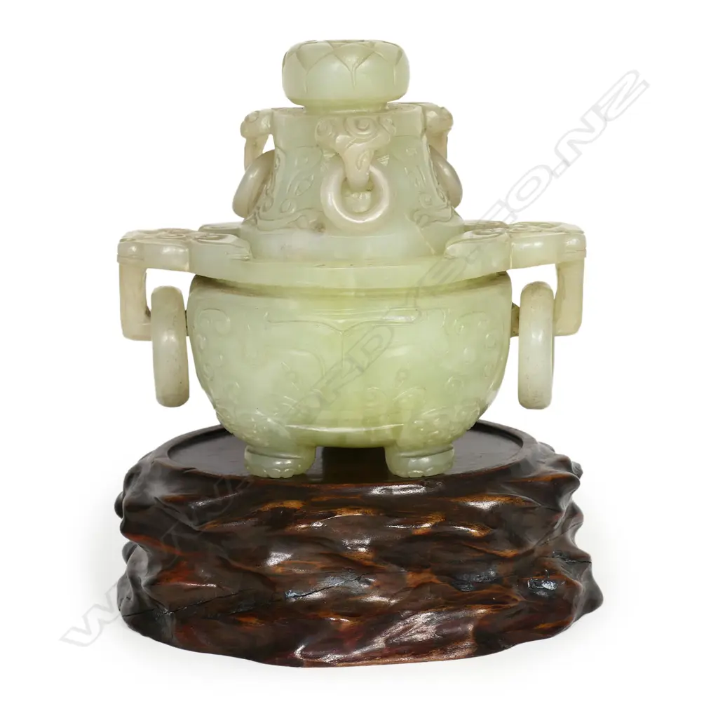 An old Chinese green jade lidded censor - the lid with pronounced flower head finial and three captured ring handles. carved with dragon masks, the body with further conforming masks to the large flat top captured ring handles. The body with opposing stylised dragons and paw feet. L.160mm. Raised on wood stand. H.195mm. Image 1++