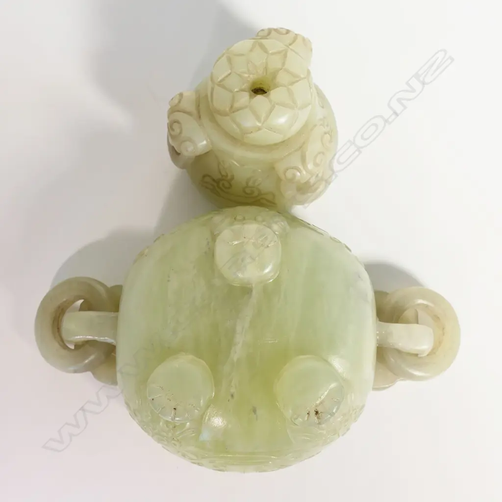 An old Chinese green jade lidded censor - the lid with pronounced flower head finial and three captured ring handles. carved with dragon masks, the body with further conforming masks to the large flat top captured ring handles. The body with opposing stylised dragons and paw feet. L.160mm. Raised on wood stand. H.195mm. Image 1++