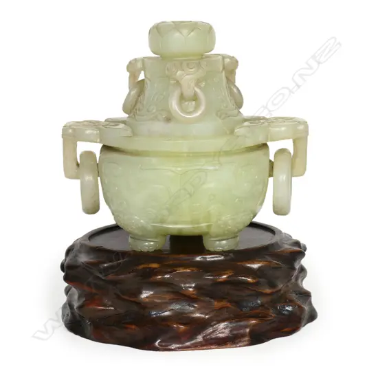 An old Chinese green jade lidded censor - the lid with pronounced flower head finial and three captured ring handles. carved with dragon masks, the body with further conforming masks to the large flat top captured ring handles. The body with opposing stylised dragons and paw feet. L.160mm. Raised on wood stand. H.195mm.