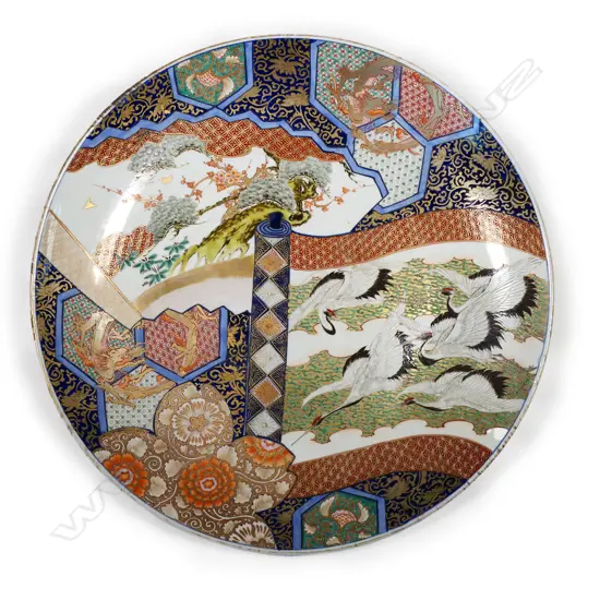 One of a matched pair, a massive Japanese Imari porcelain charger - decorated with madoe panels of 'brocade' patterns, gilt dragon rondel motifs, ancient pines and an unfurling scroll depicting Manchurian cranes in flight between ribbons of mist on a background of jade and gold cloud forms. The reverse rim painted with chrysanthemum and blossom branches in iron red and underglaze blue, Dia.600mm.