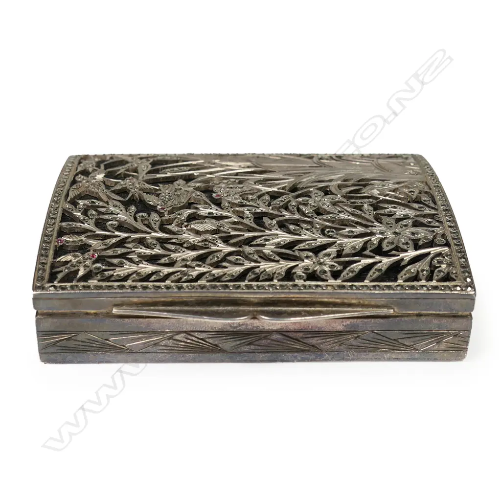 An early 20thC decorative silver box - the lid with elaborate fret cut reed, foliate and bird decorated scene jewelled with marcasite's (minor losses) and pink stones on black probably silk ground, geometric engraved Art Deco style decoration. 80 x 60 x 15mm. 118gms. Image 1++