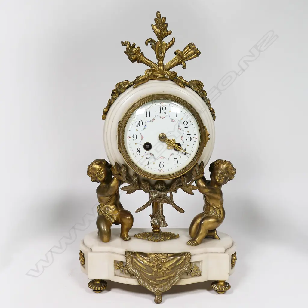 A Louis Phillipe period French marble and ormolu cased three-piece clock garniture - the clock with bell striking movement in a cream marble drum case supported by ormolu putto and surmounted by a military trophy; raised on a plateau with further gilt bronze embellishment. With a pair of associated urn-candlesticks with ram's head handles and with reversible candlestick nozzles/knopped finials. Clock H.350mm, Image 1++