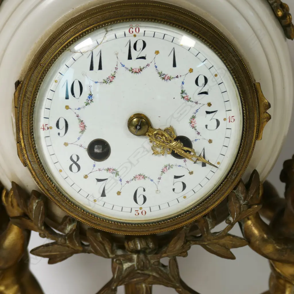 A Louis Phillipe period French marble and ormolu cased three-piece clock garniture - the clock with bell striking movement in a cream marble drum case supported by ormolu putto and surmounted by a military trophy; raised on a plateau with further gilt bronze embellishment. With a pair of associated urn-candlesticks with ram's head handles and with reversible candlestick nozzles/knopped finials. Clock H.350mm, Image 1++