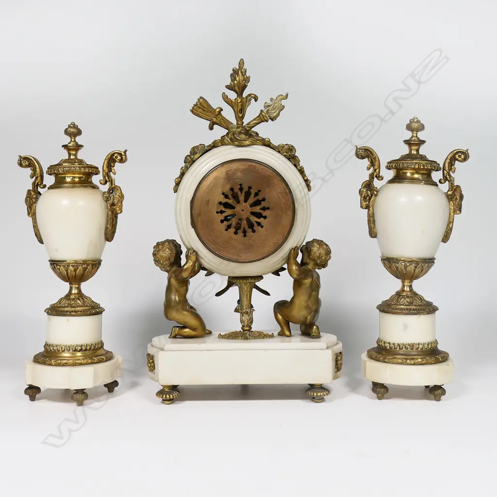 A Louis Phillipe period French marble and ormolu cased three-piece clock garniture - the clock with bell striking movement in a cream marble drum case supported by ormolu putto and surmounted by a military trophy; raised on a plateau with further gilt bronze embellishment. With a pair of associated urn-candlesticks with ram's head handles and with reversible candlestick nozzles/knopped finials. Clock H.350mm, Image 1++