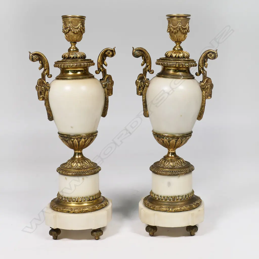 A Louis Phillipe period French marble and ormolu cased three-piece clock garniture - the clock with bell striking movement in a cream marble drum case supported by ormolu putto and surmounted by a military trophy; raised on a plateau with further gilt bronze embellishment. With a pair of associated urn-candlesticks with ram's head handles and with reversible candlestick nozzles/knopped finials. Clock H.350mm, Image 1++