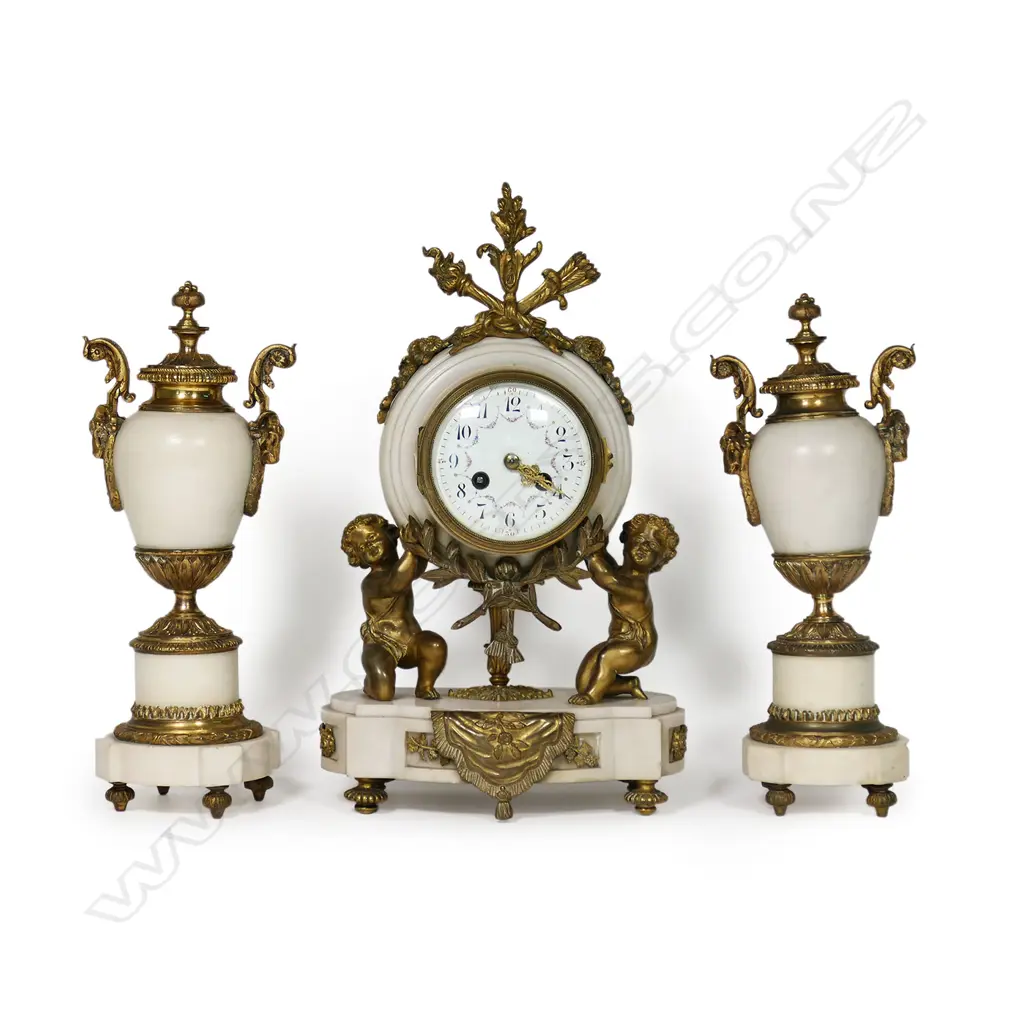 A Louis Phillipe period French marble and ormolu cased three-piece clock garniture - the clock with bell striking movement in a cream marble drum case supported by ormolu putto and surmounted by a military trophy; raised on a plateau with further gilt bronze embellishment. With a pair of associated urn-candlesticks with ram's head handles and with reversible candlestick nozzles/knopped finials. Clock H.350mm, Image 1++