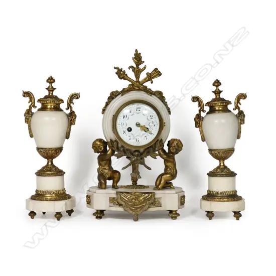 A Louis Phillipe period French marble and ormolu cased three-piece clock garniture - the clock with bell striking movement in a cream marble drum case supported by ormolu putto and surmounted by a military trophy; raised on a plateau with further gilt bronze embellishment. With a pair of associated urn-candlesticks with ram's head handles and with reversible candlestick nozzles/knopped finials. Clock H.350mm,