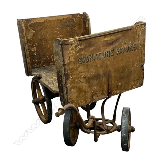 An early 1900s wooden library trolley - open sides on cast iron base and wheels, with remains of old paper and paint finish, gilt 'Signature Books' lettered to the front. 1400 x 540 x 800mm.