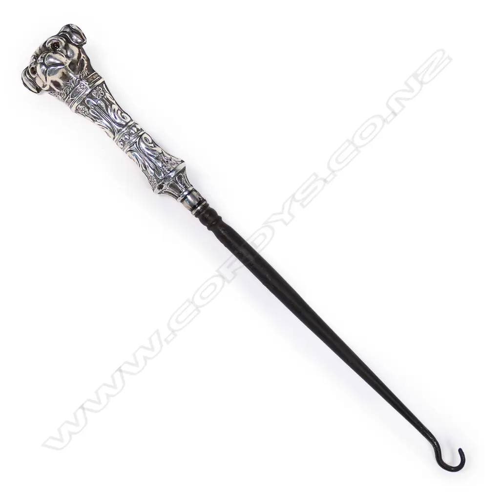 An uncommon Edwardian silver dog handled boot hook, the pug type dog with glass eyes, with collar and leafy and floral bands, steel shaft. L.230mm. Birmingham 1907, by Crisford & Norris Ltd. Minor faults consistent with age. Image 1++