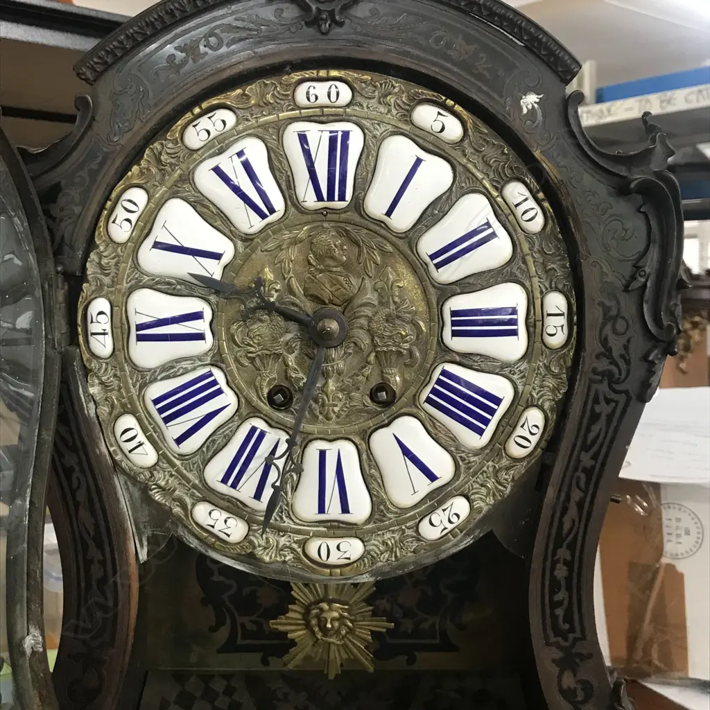 A Louis XVI-style mantel clock, the shaped case inlaid with cut brass Boulle work on black lacquered ground and heavily tarnished rococo ormolu mounts, surmounted by a pagoda top crowned by a standing heraldic angel figure. The cast dial with white enamel hour cartouches with blue Roman numerals, gong striking movement, sunburst pendulum above a sloping checkered floor, behind a glazed door mounted with a floral adorned putti. H.640mm. Image 1++