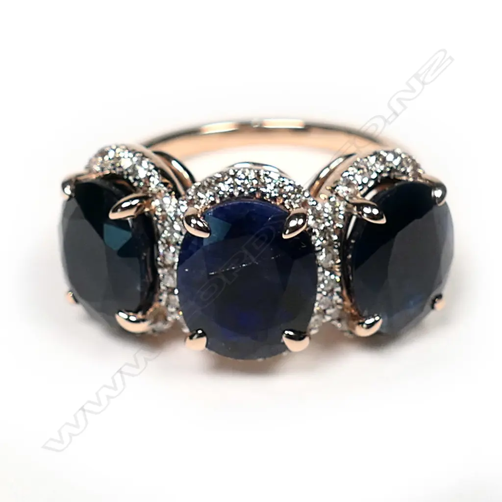 A 14ct rose gold sapphire and diamond halo trilogy ring, three oval cut dark blue sapphires (slightly included), claw set, each sapphire surrounded by a halo of round brilliant cut diamonds (J-M colour, SI1-P1 clarity), TSW 9.31ct, TDW 0.55ct, Ring Size O1/2. Image 1++