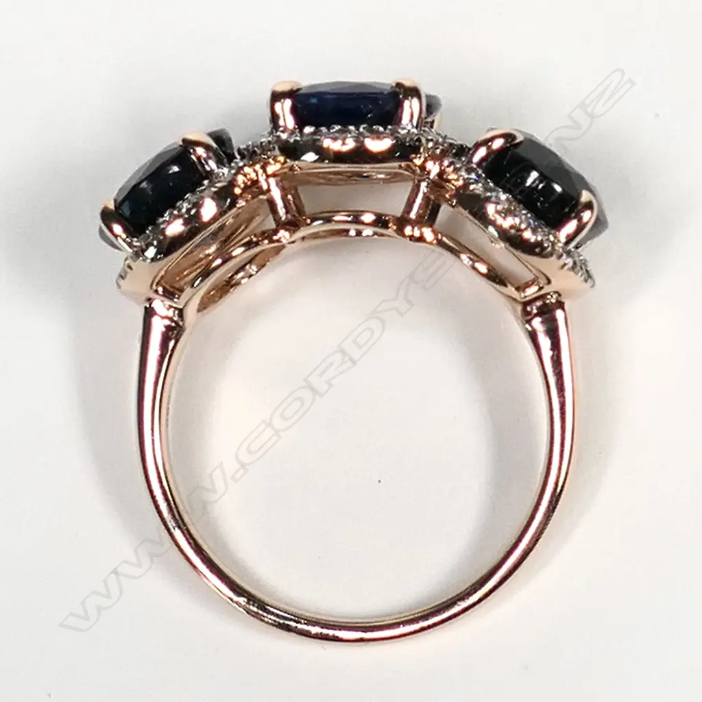 A 14ct rose gold sapphire and diamond halo trilogy ring, three oval cut dark blue sapphires (slightly included), claw set, each sapphire surrounded by a halo of round brilliant cut diamonds (J-M colour, SI1-P1 clarity), TSW 9.31ct, TDW 0.55ct, Ring Size O1/2. Image 1++