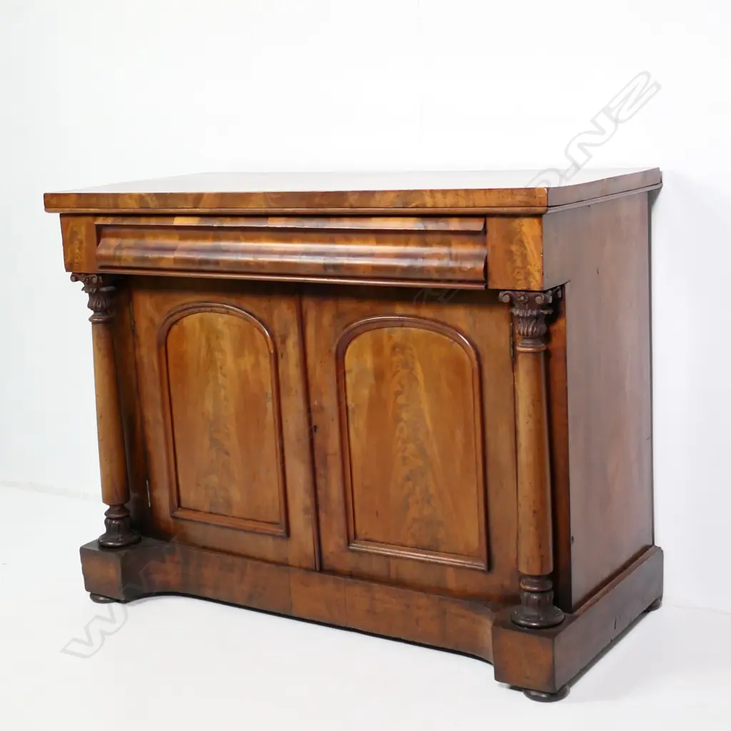 A Regency flame mahogany small sideboard/chiffonier, scrolling back, frieze drawer to the top with full Corinthian columns to each side of two panelled doors, raised on plinth base with squat bun feet.  (Sun fading to one side) 1230 x 575 x 1520mm. Note: This piece does have a scroll back, not shown in photo. Image 1++