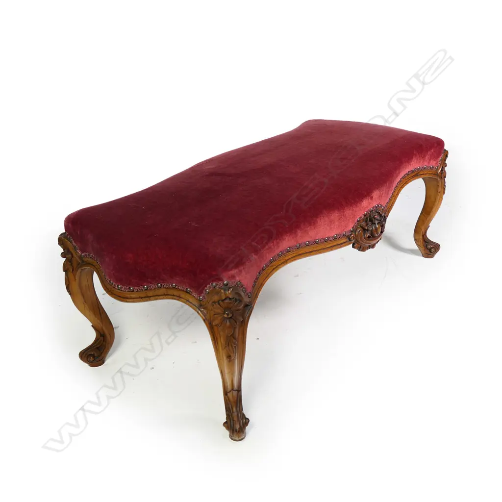A Victorian walnut footstool, upholstered in a maroon velvet, the cabriole legs with floral knees, fruit and floral decoration to the centre of the frame.  1120 x 530 x 420mm. Image 1++