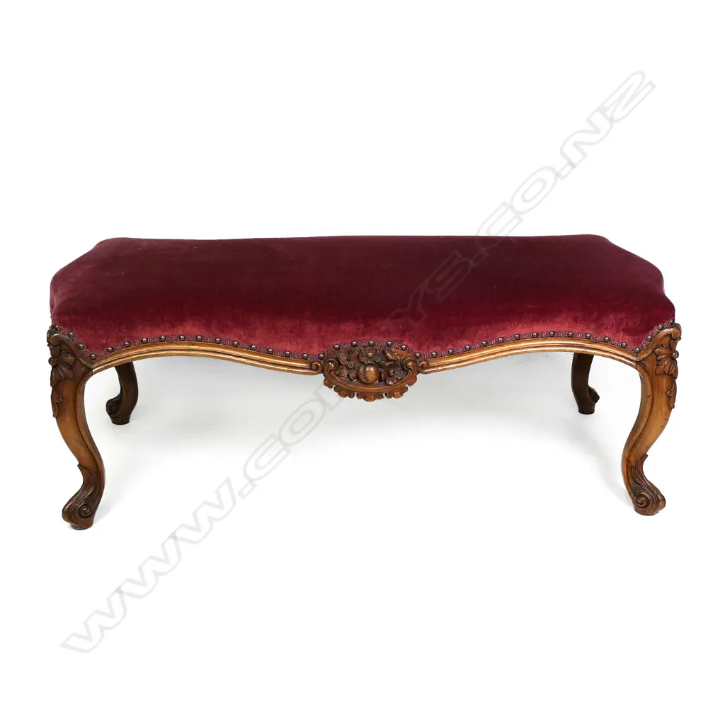 A Victorian walnut footstool, upholstered in a maroon velvet, the cabriole legs with floral knees, fruit and floral decoration to the centre of the frame.  1120 x 530 x 420mm. Image 1++