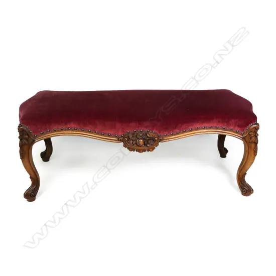 A Victorian walnut footstool, upholstered in a maroon velvet, the cabriole legs with floral knees, fruit and floral decoration to the centre of the frame.  1120 x 530 x 420mm.