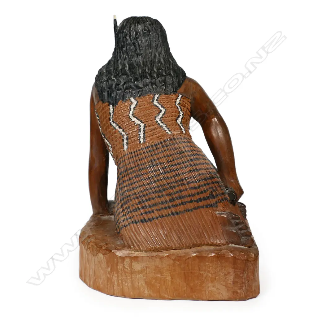 A carved folk art wahine, kneeling in traditional garments with a huia feather tucked behind her ear, and holding a patu in her right hand. H.220mm. Image 1++