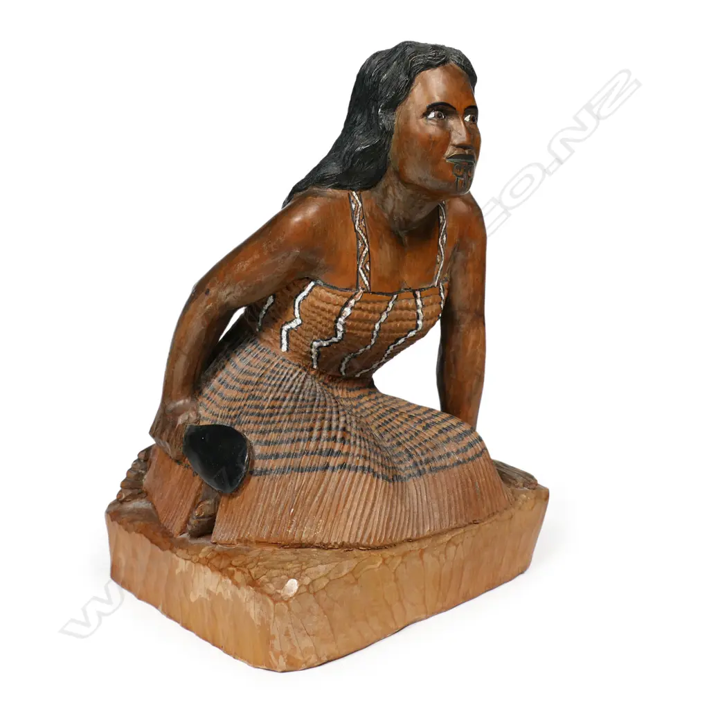 A carved folk art wahine, kneeling in traditional garments with a huia feather tucked behind her ear, and holding a patu in her right hand. H.220mm. Image 1++