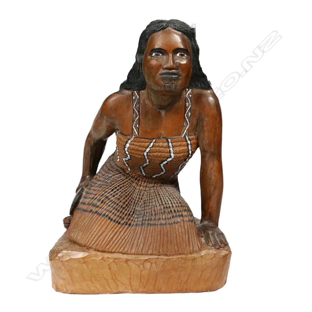 A carved folk art wahine, kneeling in traditional garments with a huia feather tucked behind her ear, and holding a patu in her right hand. H.220mm. Image 1++