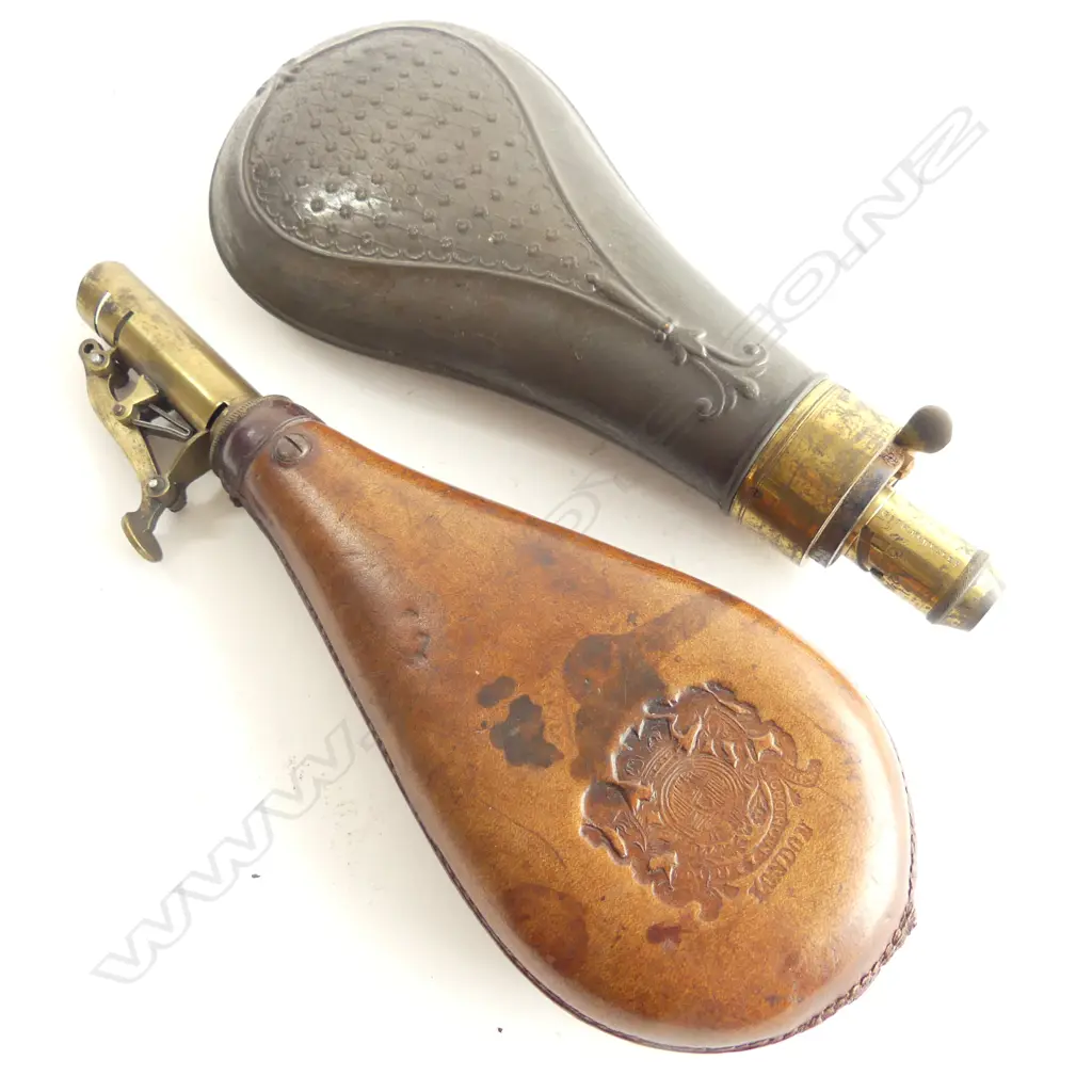 Two bag shaped powder flasks; the leather one with stamped London coat-of-arms and sprung lever brass dispenser nozzle; the other of pressed tin marked ‘G & J W Hawkes’ to the brass graduated nozzle. Image 1++