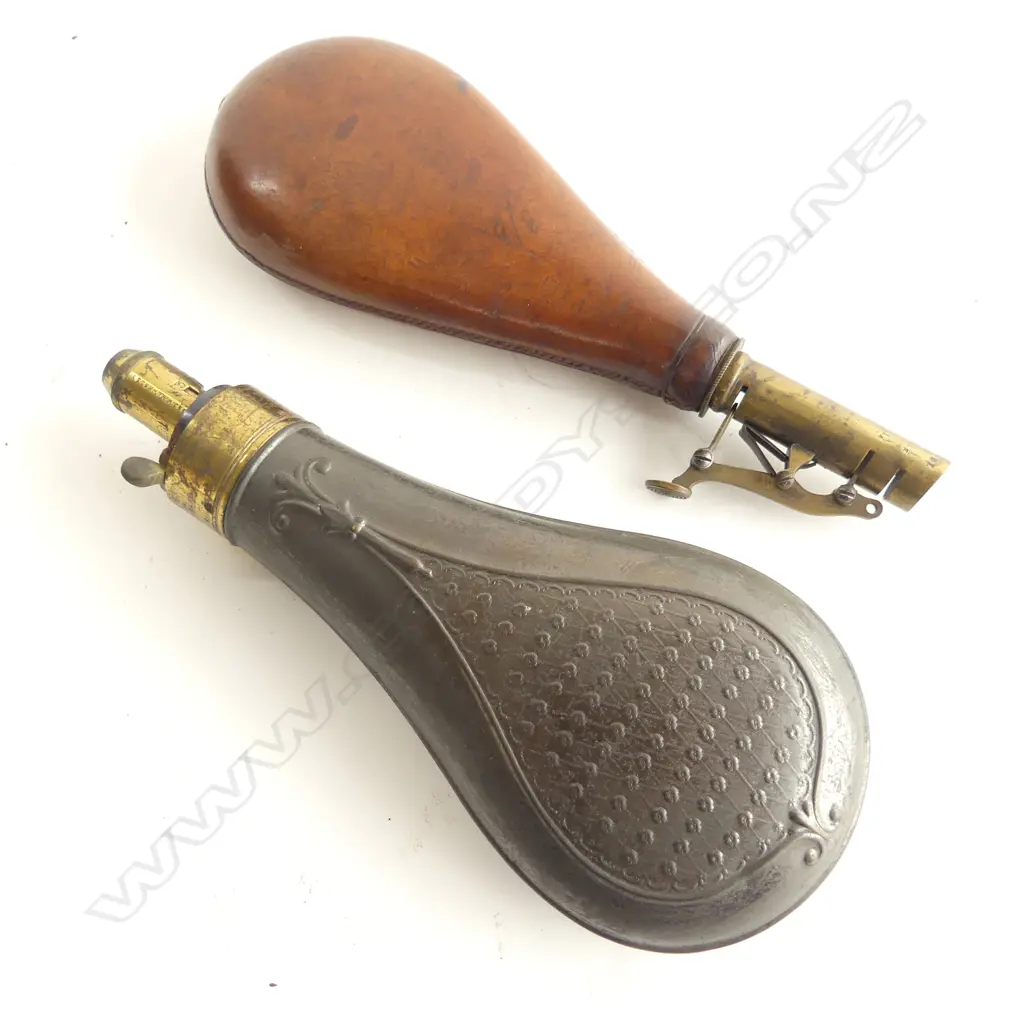 Two bag shaped powder flasks; the leather one with stamped London coat-of-arms and sprung lever brass dispenser nozzle; the other of pressed tin marked ‘G & J W Hawkes’ to the brass graduated nozzle. Image 1++